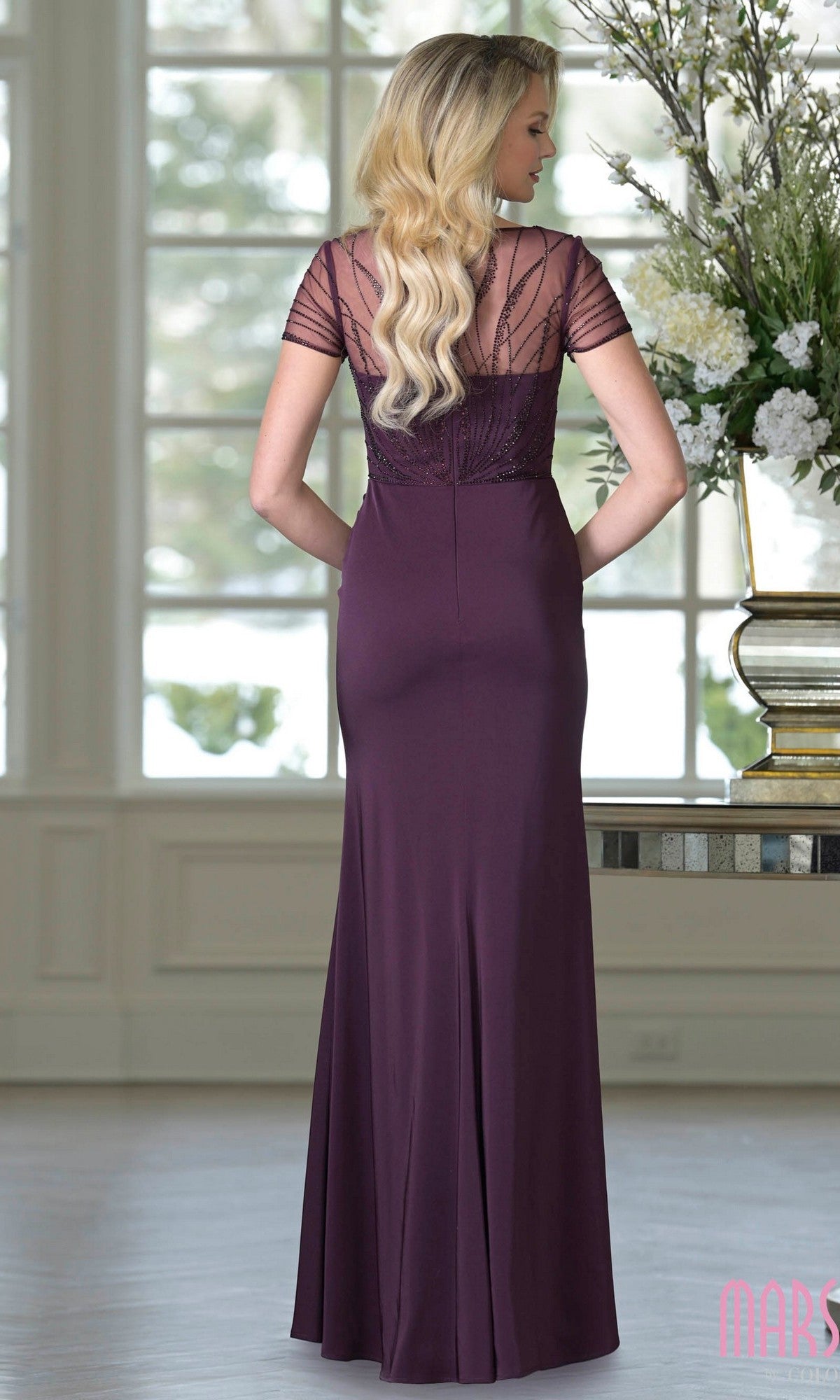 A woman with long, wavy blonde hair wears the Marsoni M350, a floor-length plum formal dress with short sleeves, lace detailing, and a beaded bodice, standing indoors and facing away from the camera.